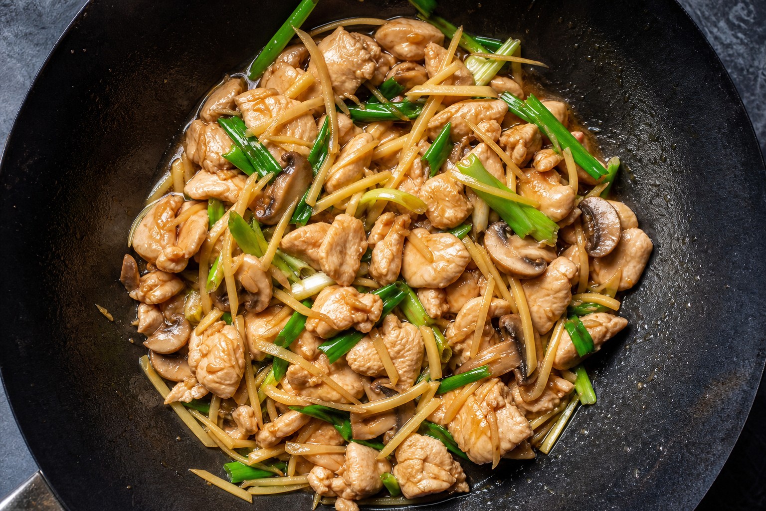 Chicken and ginger stir fry with mushrooms and spring onions, in a black wok seen from above