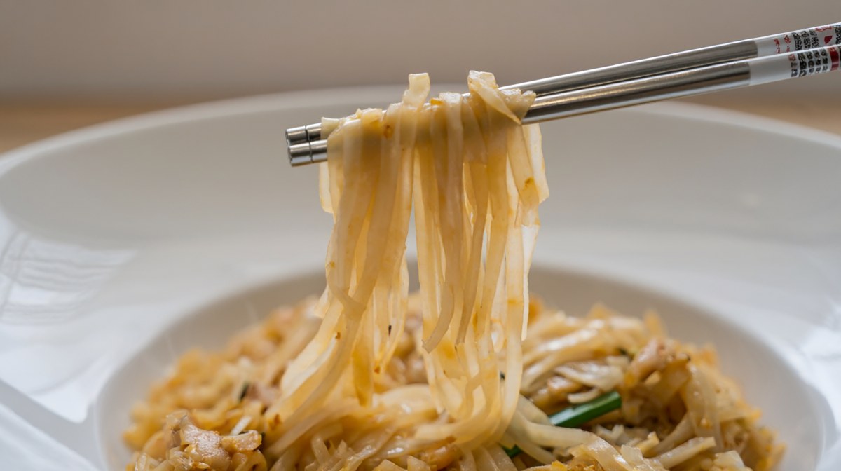 Chopsticks lifting pad thai from a bowl, the noodles hanging in a soft even drape