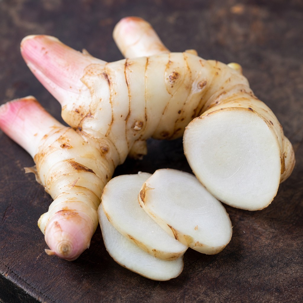 Fresh galangal root on a dark wooden board — knobbly pale cream skin with pink-tinged tips