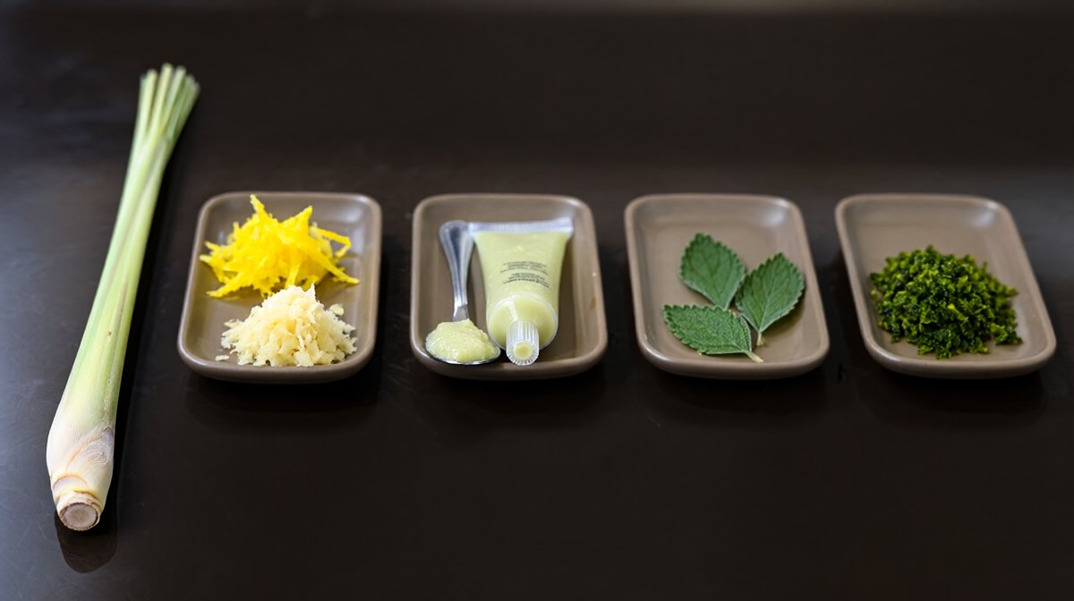 A fresh lemongrass stalk beside four small dishes in a row, lemon zest with grated ginger, a tube of pale green paste, lemon verbena leaves and kaffir lime zest