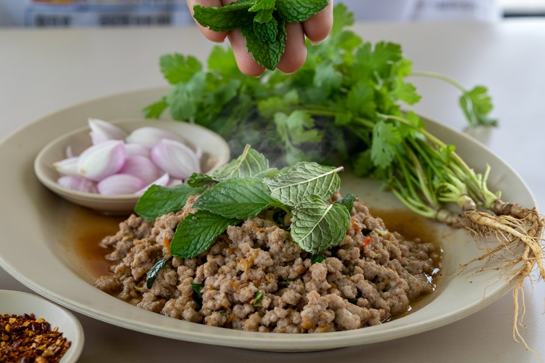 A hand scattering whole mint leaves over warm Isaan minced meat salad, with coriander, paper-thin sliced shallots and dried chilli flakes beside the plate