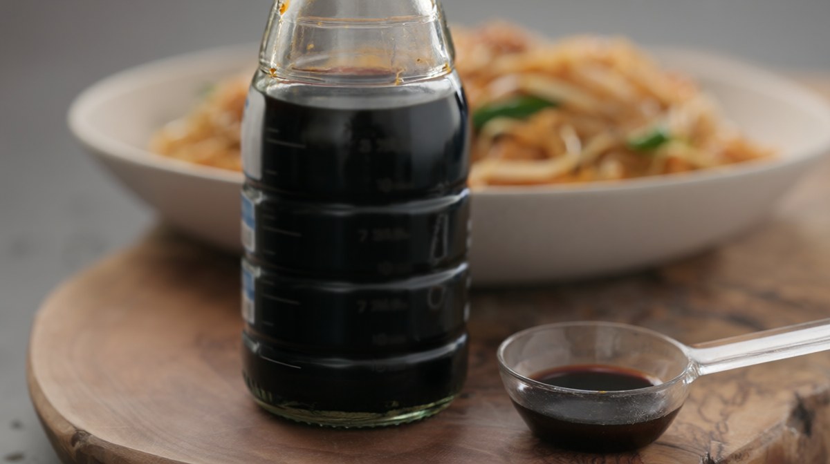 A plain bottle of fish sauce and a measuring spoon beside a bowl of pad thai in the background