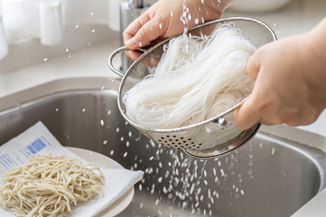 Hands shaking a colander of soaked rice noodles hard over a sink with water spraying off them, kitchen paper and a plate of dry noodles beside it